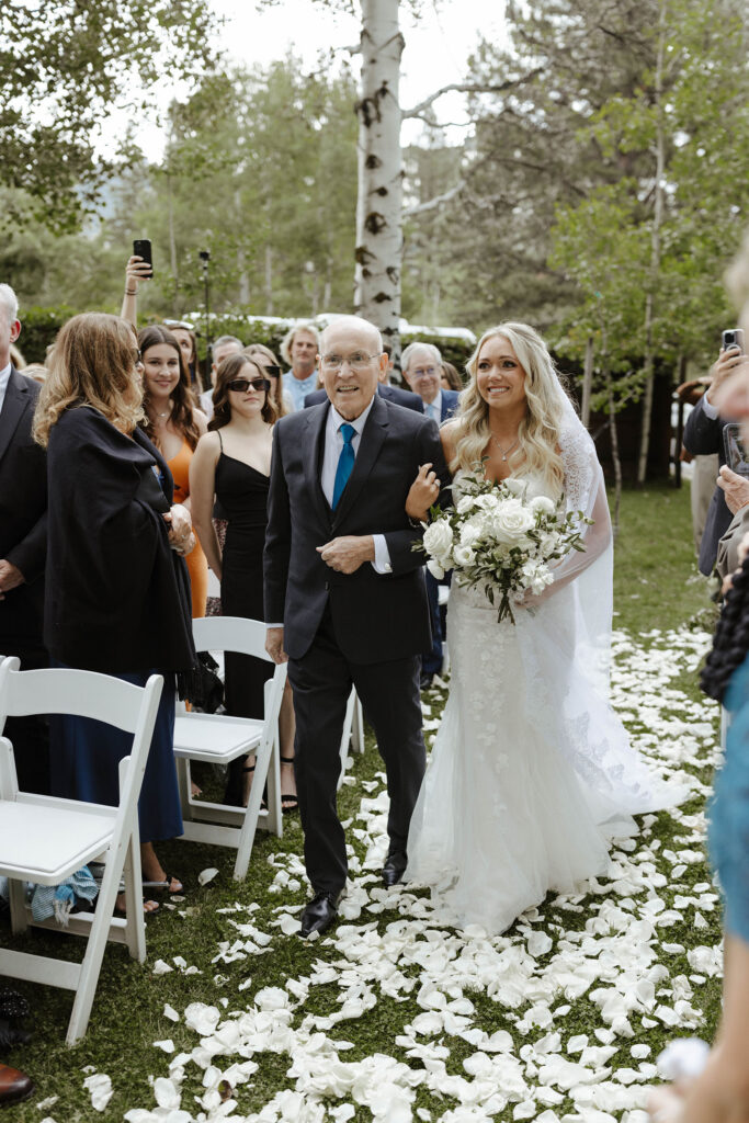 Bride smiling while walking down wedding aisle on white flower petals while holding dad's arm at Aspen Grove