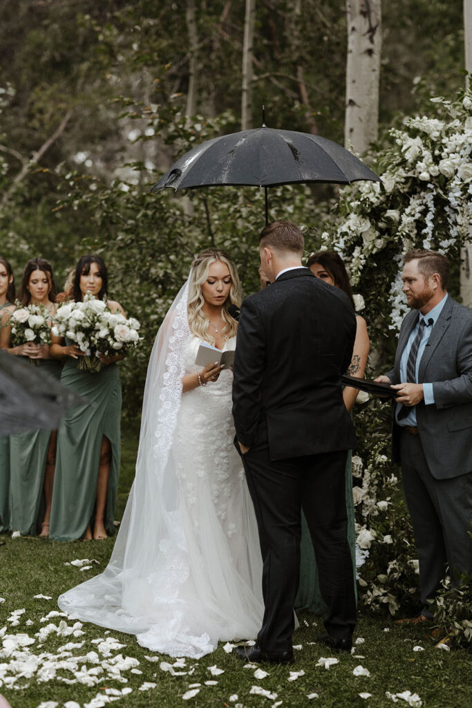 Groom holding umbrella while bride reads vows during wedding ceremony in the rain at Aspen Grove