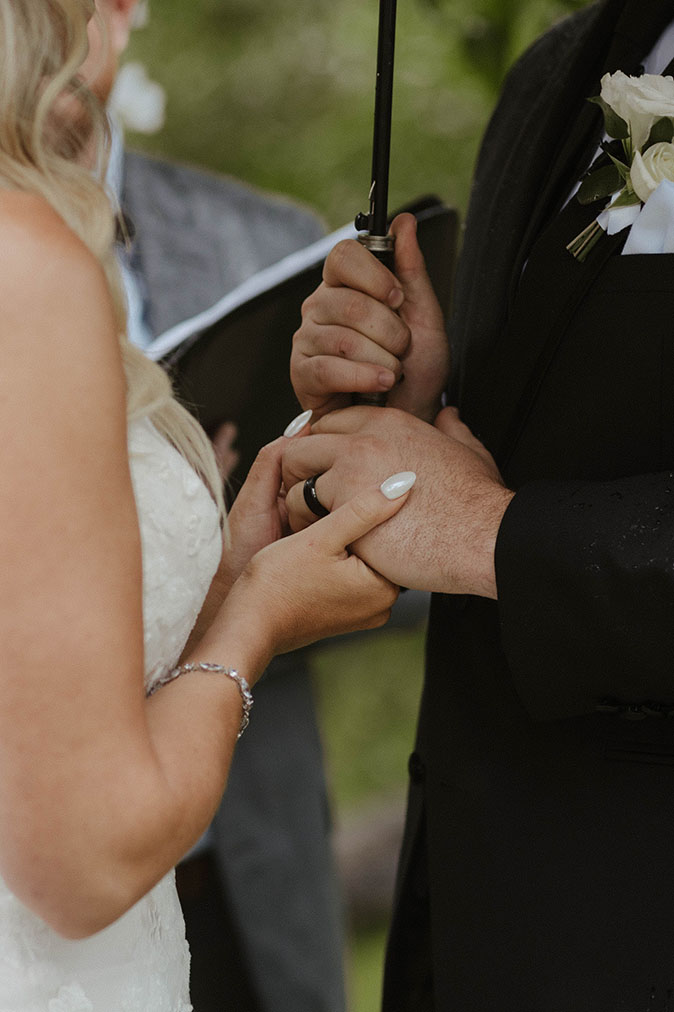 Close up of bride holding grooms hand with wedding ring while groom holds umbrella with other hand during ceremony at Aspen Grove