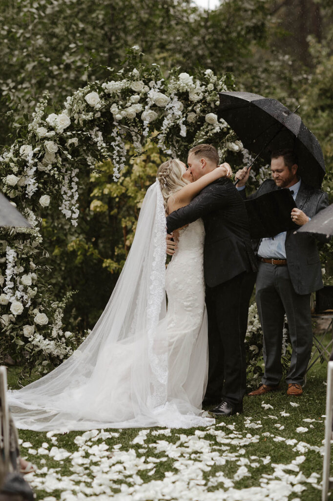 Wedding couple kissing after ceremony in front of arch with hanging white flowers and petals on the ground in front of them at Aspen Grove