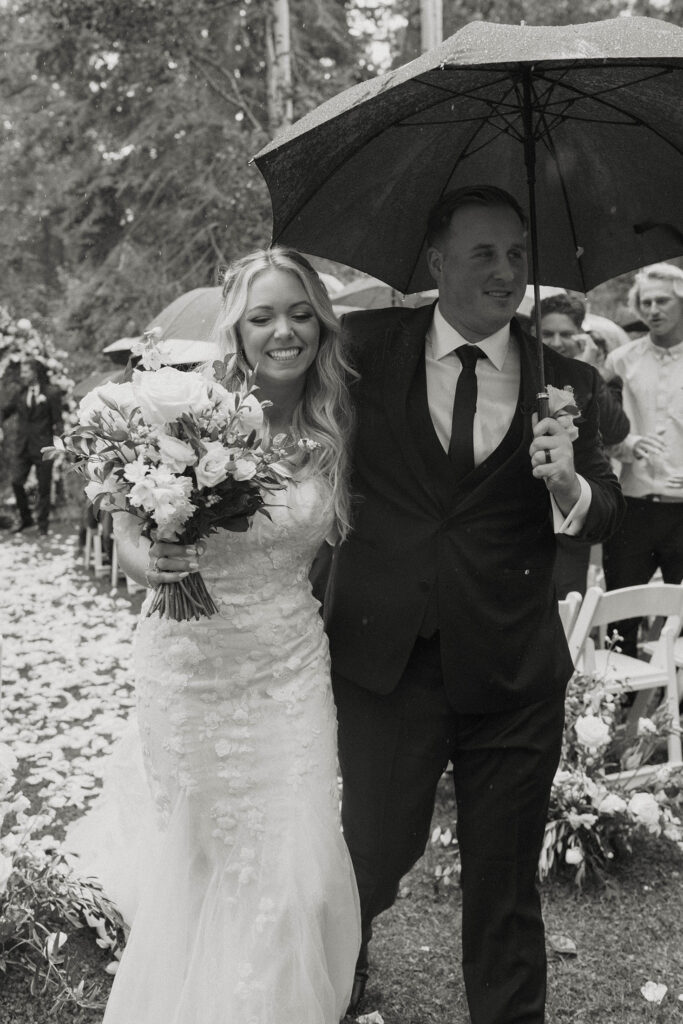 Wedding couple walking down aisle together after ceremony smiling while bride holds bouquet and groom holds umbrella at Aspen Grove