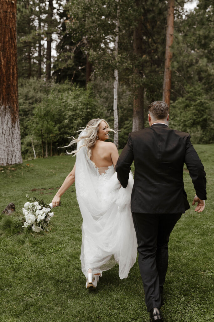 Wedding bride smiling while running and looking back over shoulder at groom on grass with trees in background at Aspen Grove
