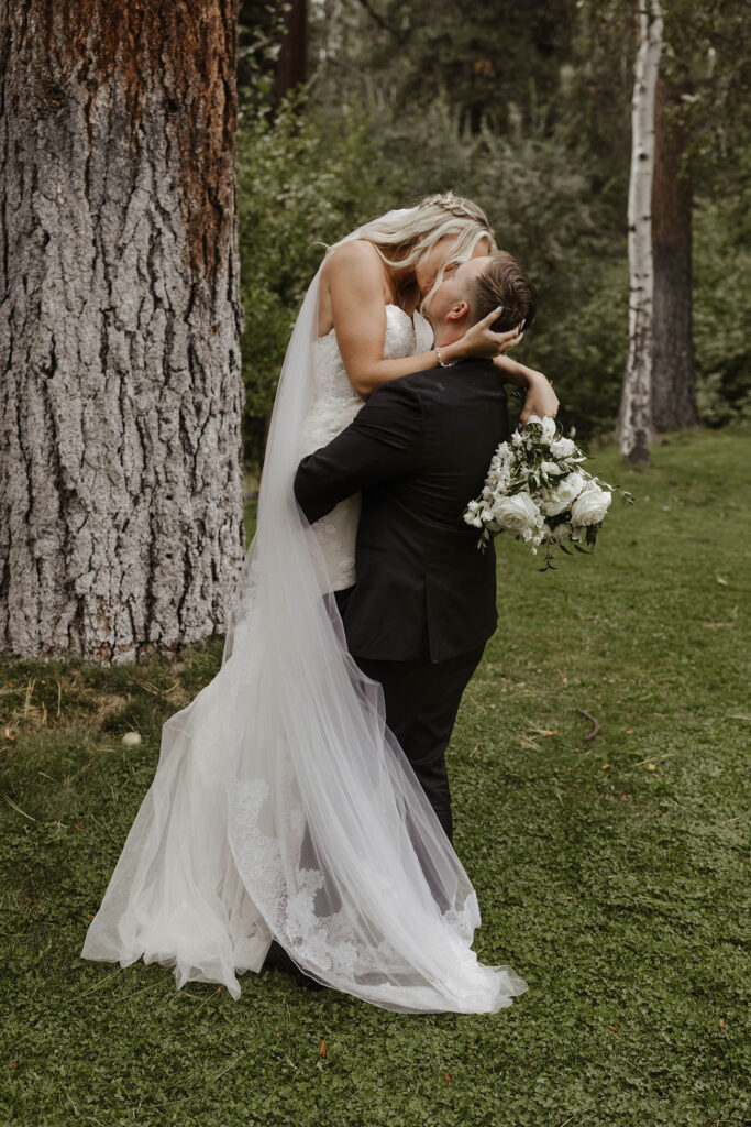 Groom lifting up and kissing bride while on grass at Aspen Grove with trees in background