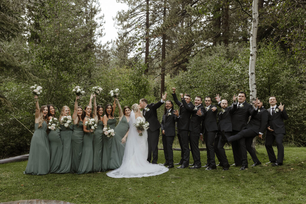 Wedding couple kissing while groomsmen and bridesmaids stand on either side and celebrate while on grass at Aspen Grove