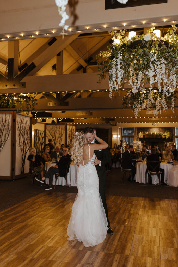 Wedding couple holding each other during first dance while guests watch with hanging floral light fixtures inside Aspen Grove