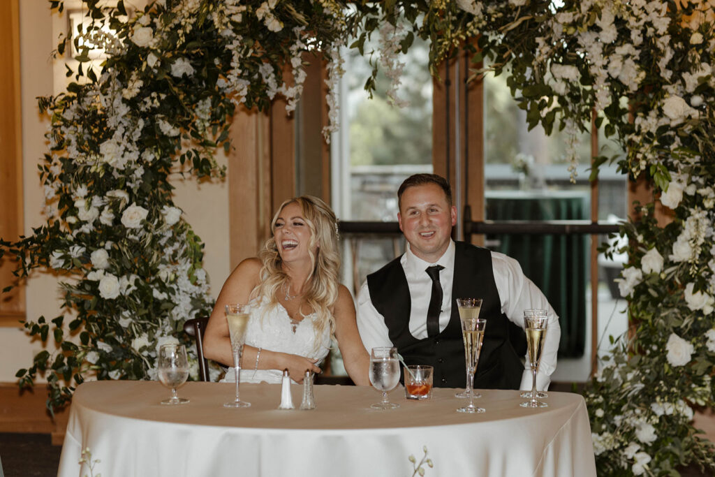 Wedding couple smiling and laughing at reception table together with arch behind them at Aspen Grove