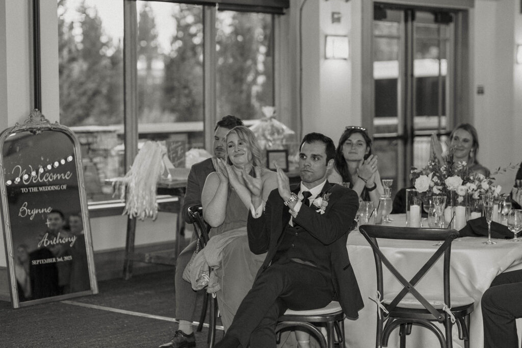 Wedding guests clapping during speeches inside at Aspen Grove with tall windows behind them