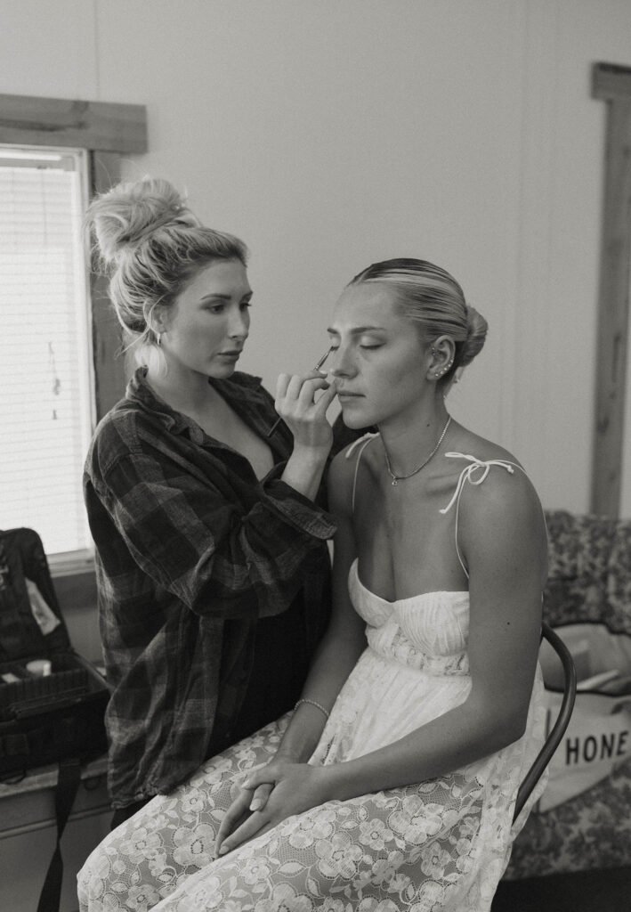 bride in her white lace pajamas having her makeup done by a makeup artist at kinship ranch