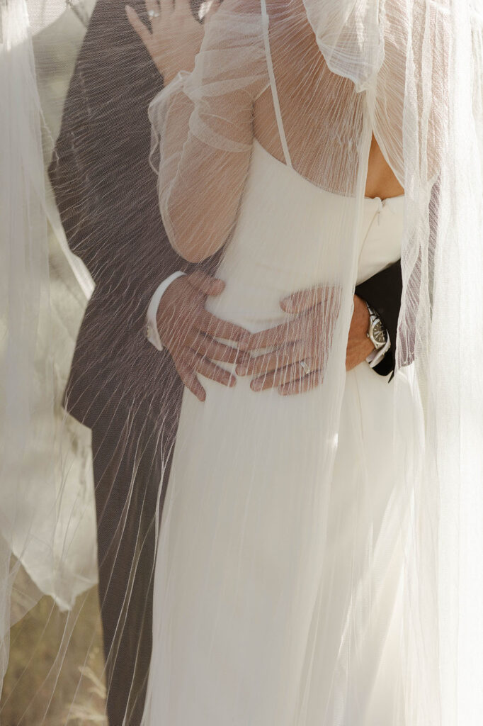 grooms hands holding the brides back with a veil covering them at a kinship ranch wedding