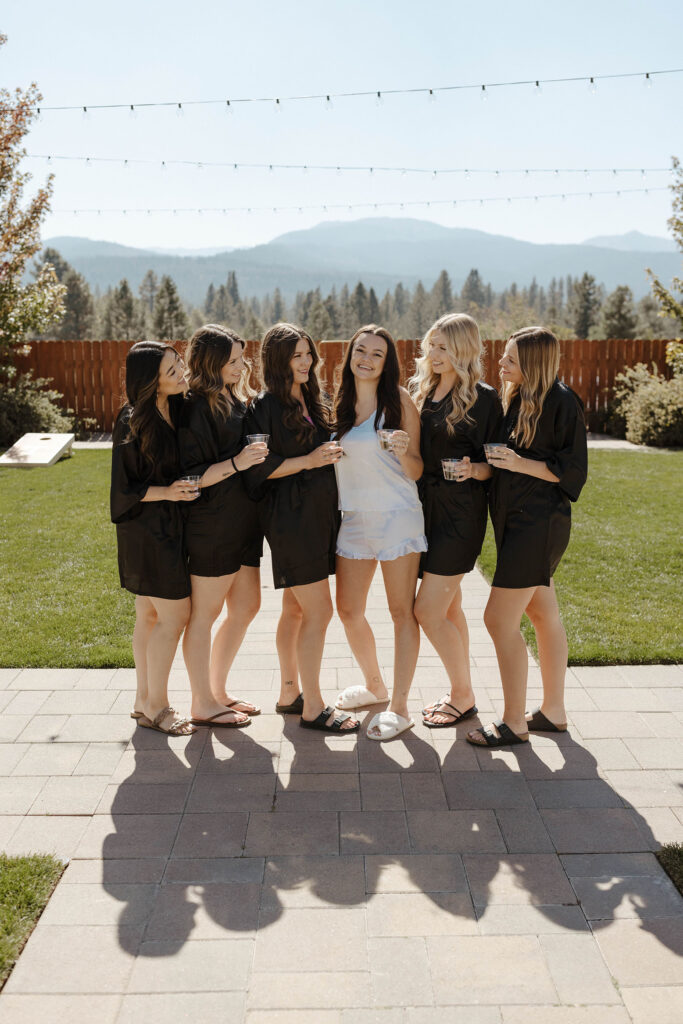 Bride standing and smiling with bridesmaids as they all hold drinks on stone walkway with grass around them at the Corner Barn