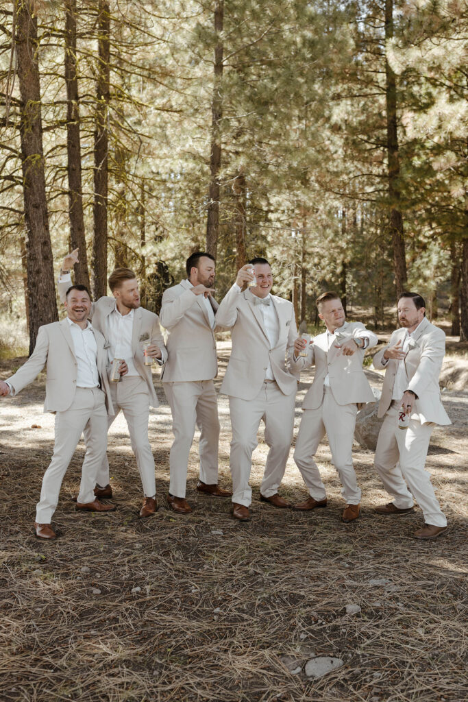 Wedding groom and groomsmen celebrating while standing together with pine trees in background at the Corner Barn
