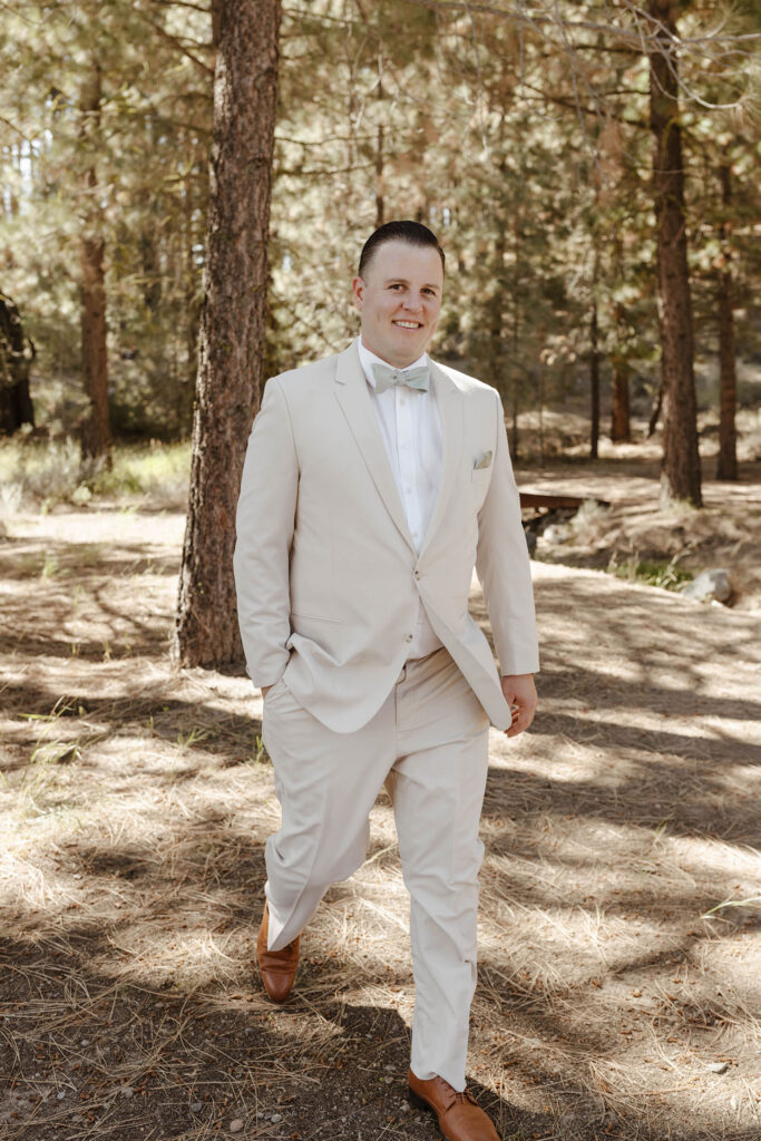 Wedding groom with hand in is pocket smiling while walking toward camera with pine trees in background at the Corner Barn