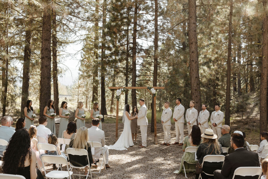 Wedding ceremony at the Corner Barn with tall pine trees in background and wooden wedding arch with white florals