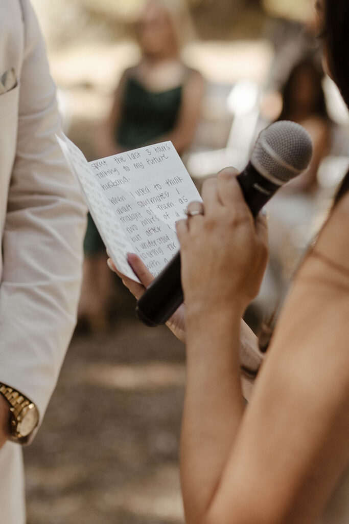 Bride holding microphone and vow book while reading wedding vows during ceremony at the Corner Barn