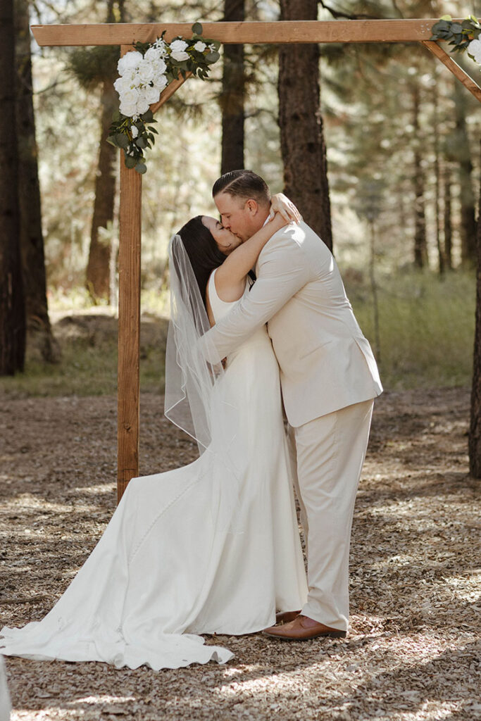 Wedding couple holding each other and kissing in front of wedding arch with white florals after ceremony at the Corner Barn