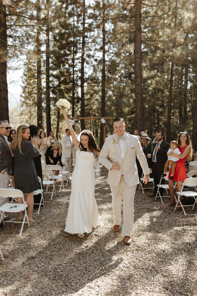 Wedding couple holding hands while walking down aisle together with bride holding up floral bouquet and guests clapping around them at the Corner Barn