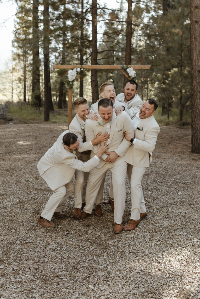 Groom laughing as groomsmen surrounded and playfully attack him with wedding arch and pine trees in background at the Corner Barn