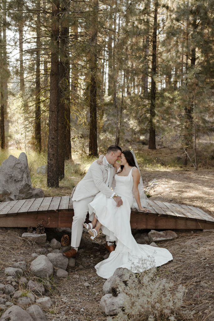 Wedding couple holding each other and touching foreheads while sitting on wooden bridge together with pine trees in background at the Corner Barn