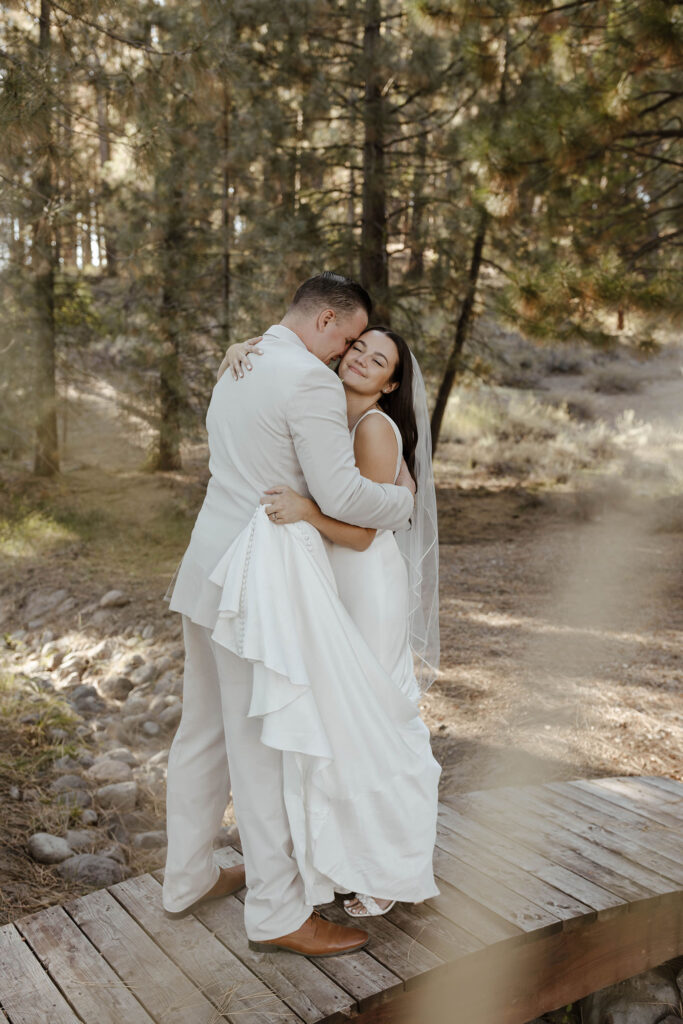Wedding bride smiling while hugging groom on wooden bridge at the Corner Barn with pine trees in background