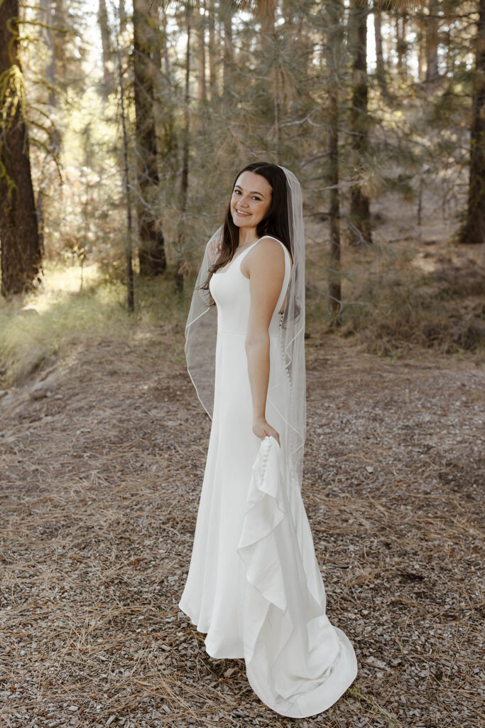 Bride holding dress tail while looking back over shoulder and smiling at camera with pine trees in background at the Corner Barn