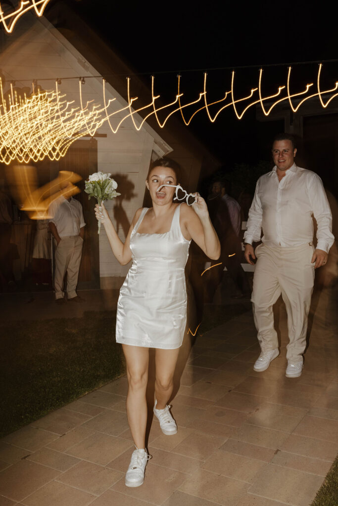 Bride dancing while holding floral bouquet and glasses with lights streaming around her and groom smiles in background at the Corner Barn