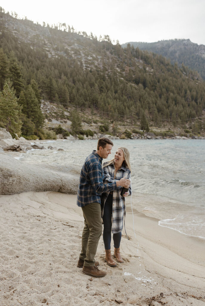 Married couple popping champagne and celebrating anniversary while on sandy beach in Lake Tahoe with pine trees in background