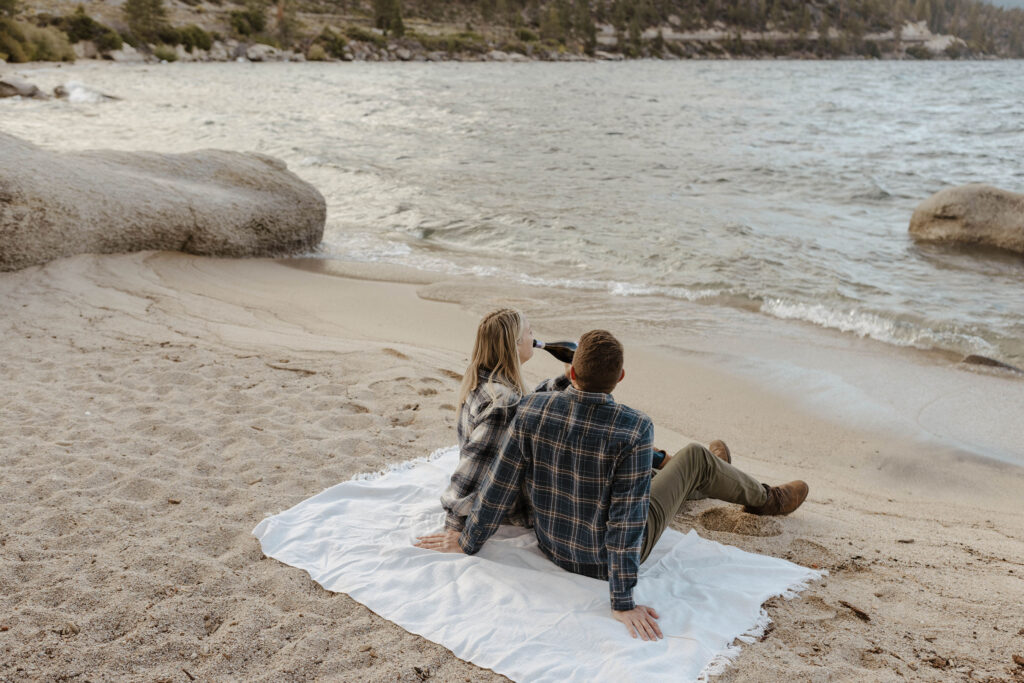 Married couple relaxing on towel together on sandy beach in Lake Tahoe as woman sips champagne bottle