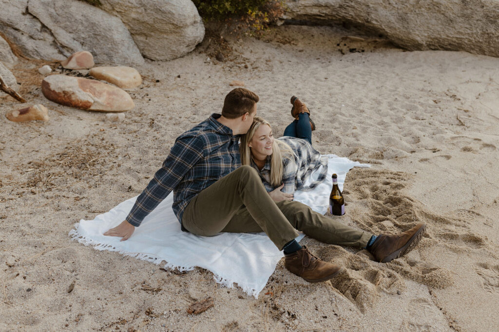 Married couple lying on towel together on sandy beach in Lake Tahoe while husband kisses wife's head