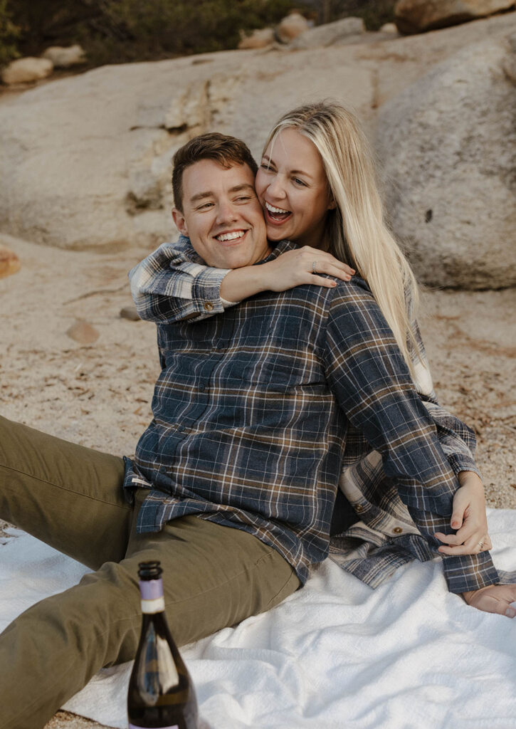 Married couple sitting on towel together on sandy beach as wife hugs husband from behind and both smile in Lake Tahoe