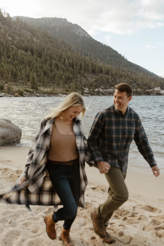 Married couple holding hands and running along sandy beach together in Lake Tahoe while husband smiles at wife with pine trees in background