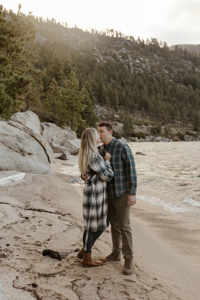 Married couple kissing and holding each other while standing on sandy beach in Lake Tahoe with rocks and trees around them