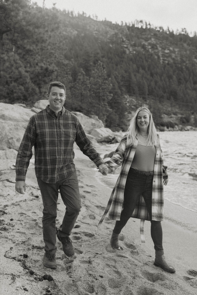 Married couple holding hands while walking toward camera and smiling on sandy beach in Lake Tahoe
