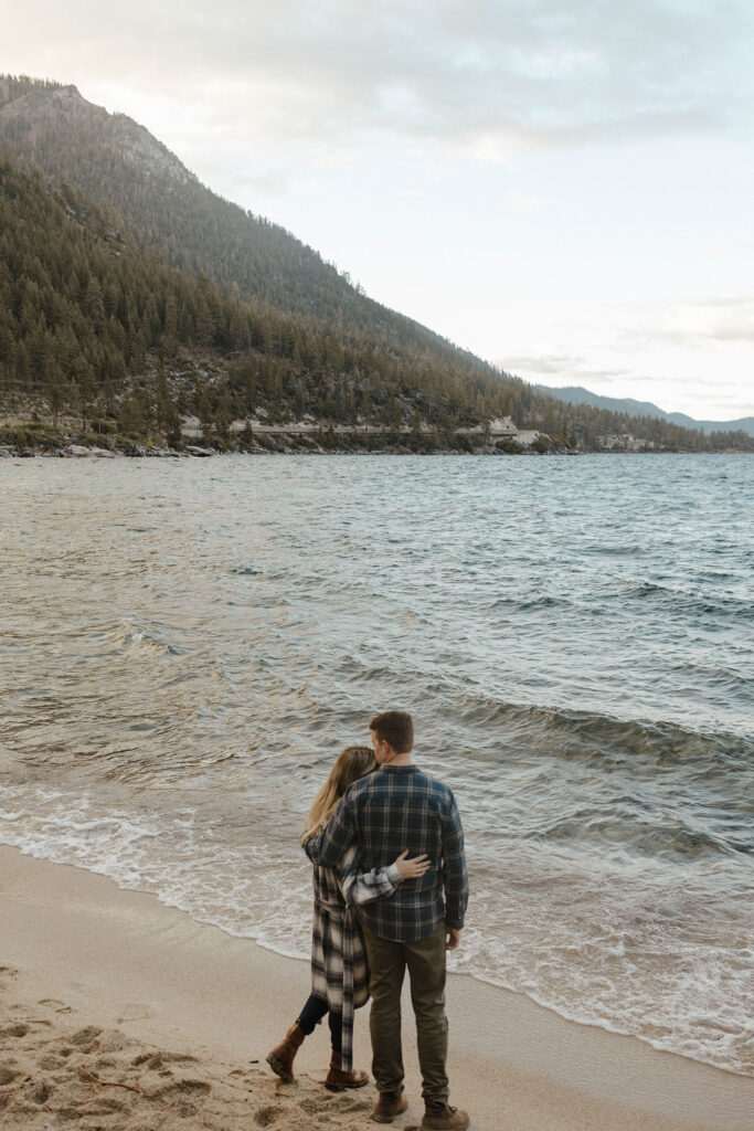 Married couple standing at the edge of Lake Tahoe looking out over the water with arms wrapped around each other