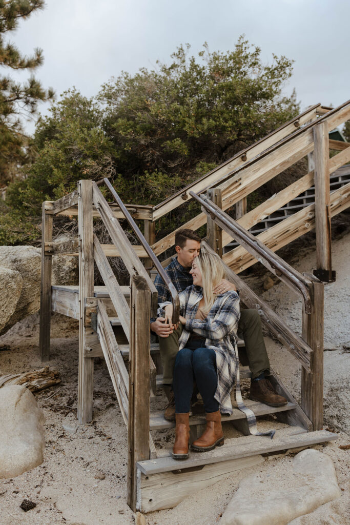 Married couple in flannels sitting on wooden stairs while hugging each other and man kisses wife's forehead on sandy beach in Lake Tahoe
