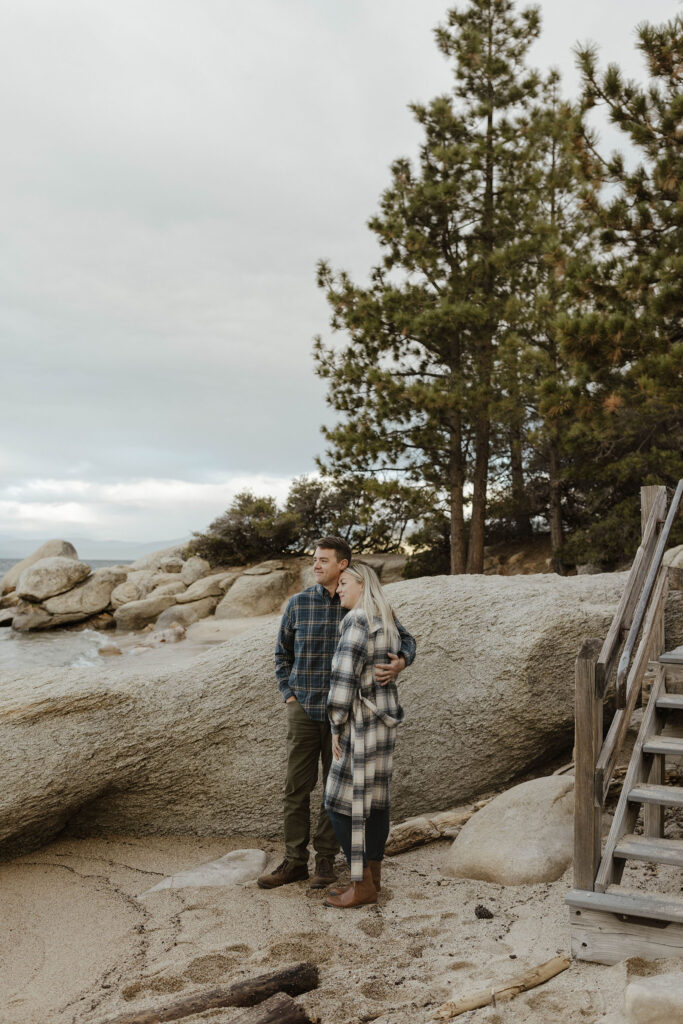 Man with arm around wife while they both look out to Lake Tahoe while standing on sandy beach with large rock behind them