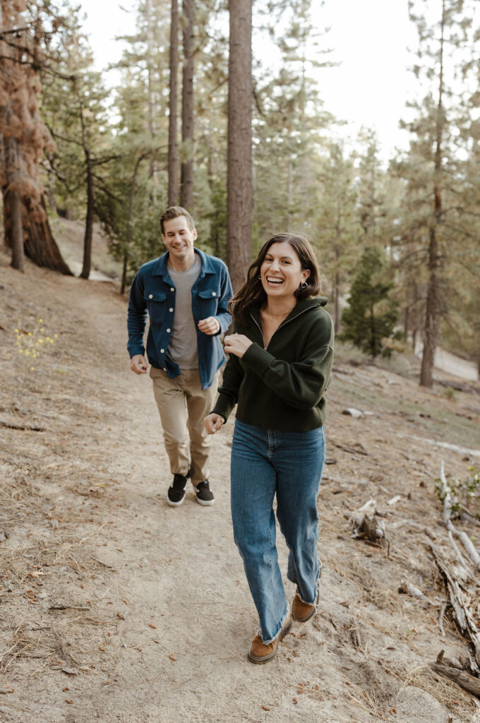 Woman laughing and looking at camera while running away from fiancé on dirt trail in Lake Tahoe