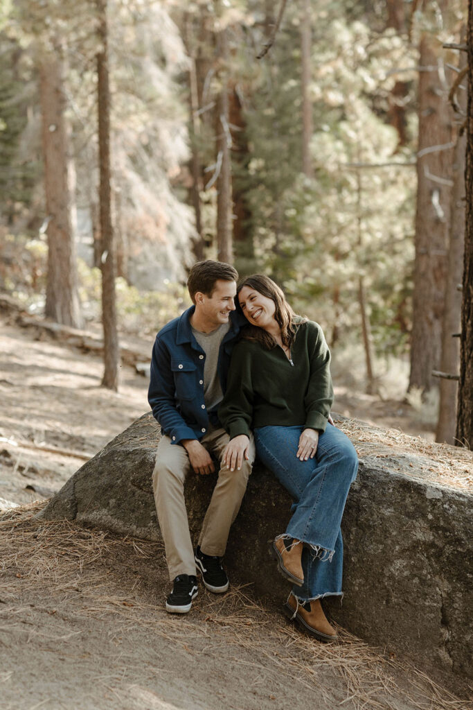 Engagement couple sitting on rock together and smiling at each other with pine trees in background at Lake Tahoe