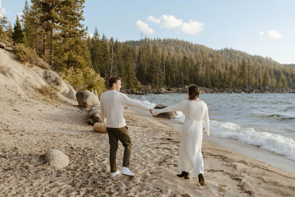 Engagement couple smiling at each other while holding outstretched hands on sandy beach in Lake Tahoe