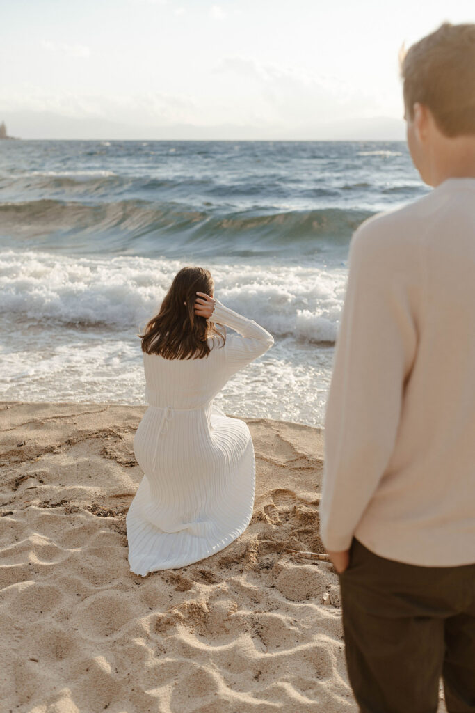 Man watching fiancé crouch by the water on a beach in Lake Tahoe with waves crashing