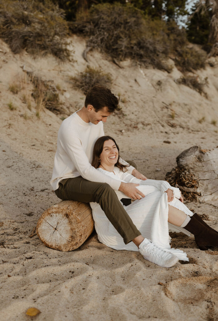 Woman leaning against fiancé while he sits on log and holds her from behind on a beach in Lake Tahoe