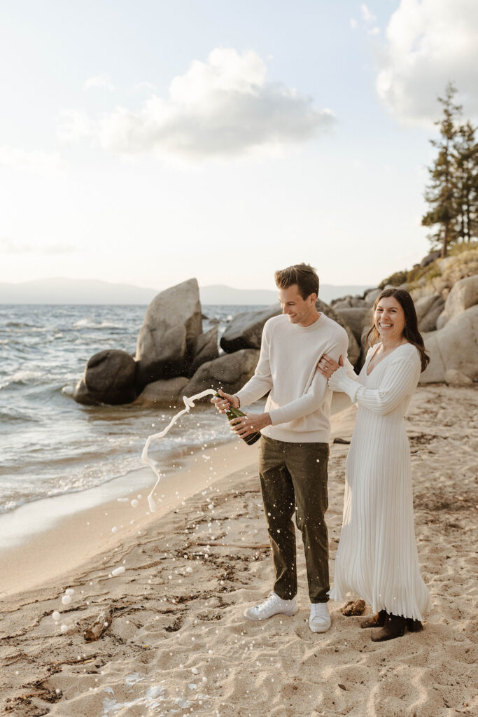 Engagement couple popping champagne bottle while on beach in Lake Tahoe with lake and rocks in background