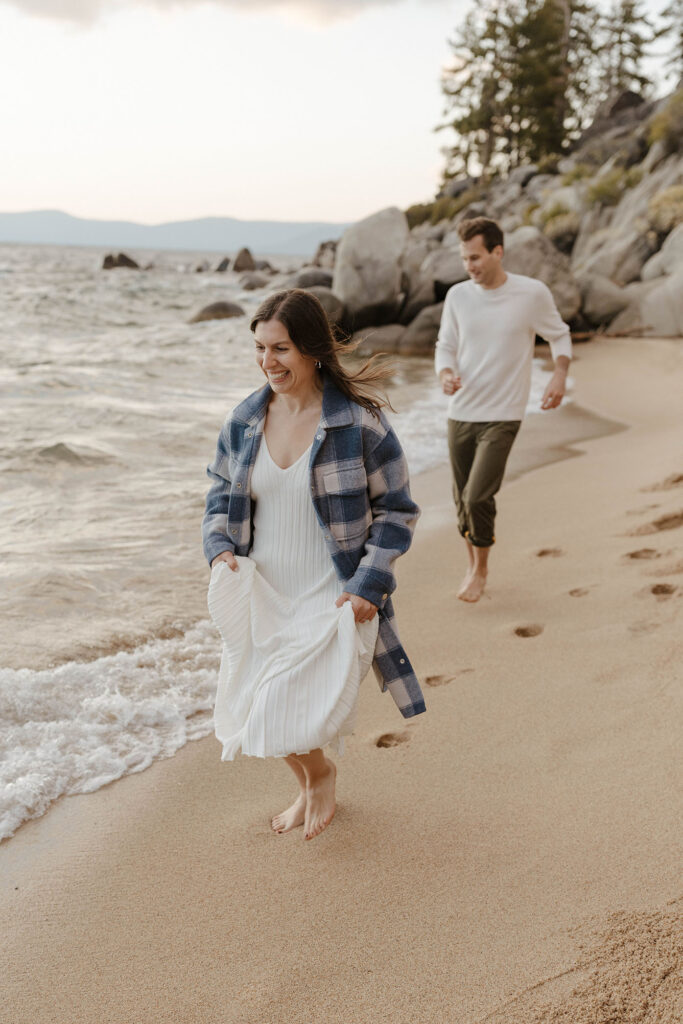 Engagement couple smiling while running across sandy beach barefoot together and woman holds her dress up in Lake Tahoe