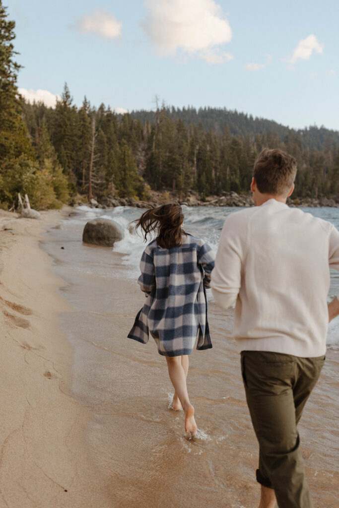 Engagement couple barefoot and running across sandy beach together in Lake Tahoe while woman wears fiancé's sweater