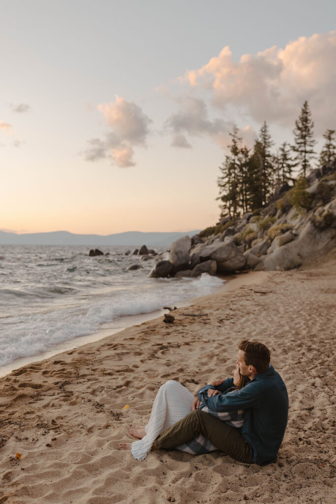 Man holding fiancé from behind while both sit on sandy beach together watching the sunset with lake and large rocks in background