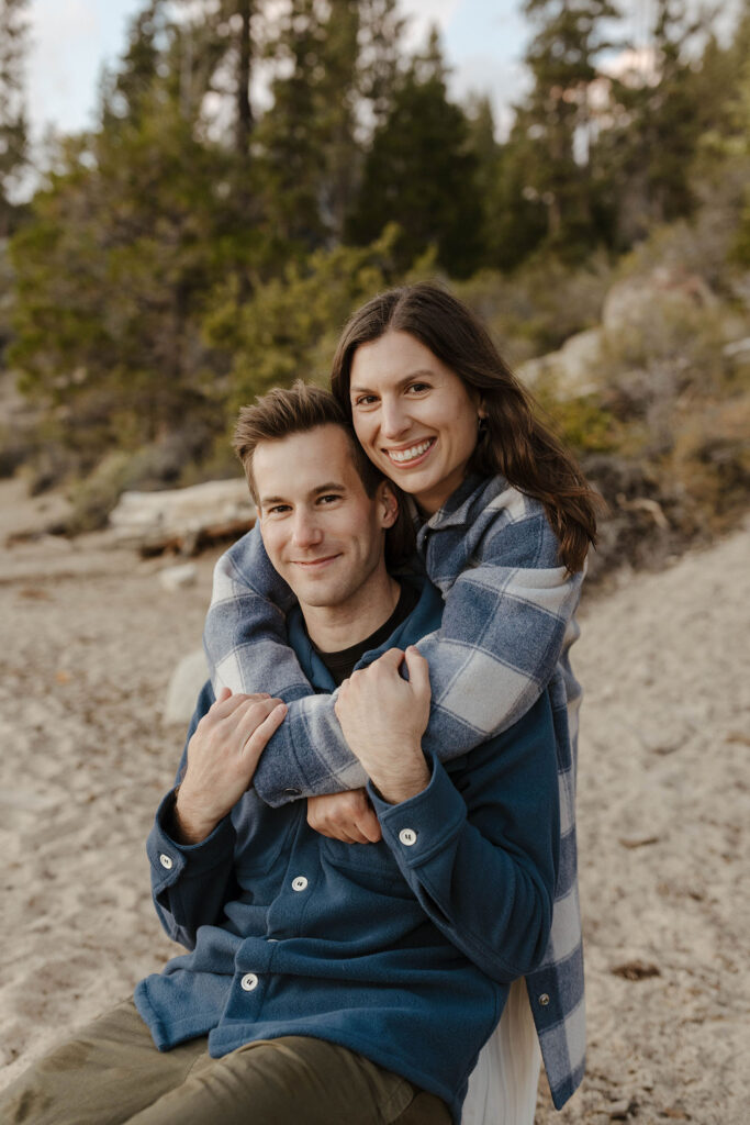 Engagement couple smiling at camera while woman hugs fiancé from behind while on beach in Lake Tahoe