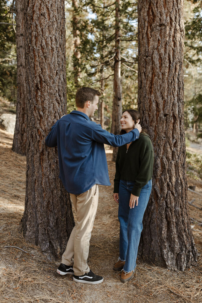 Engagement couple leaning on two tall pine trees while looking at each other and man fixes fiancé's hair in Lake Tahoe