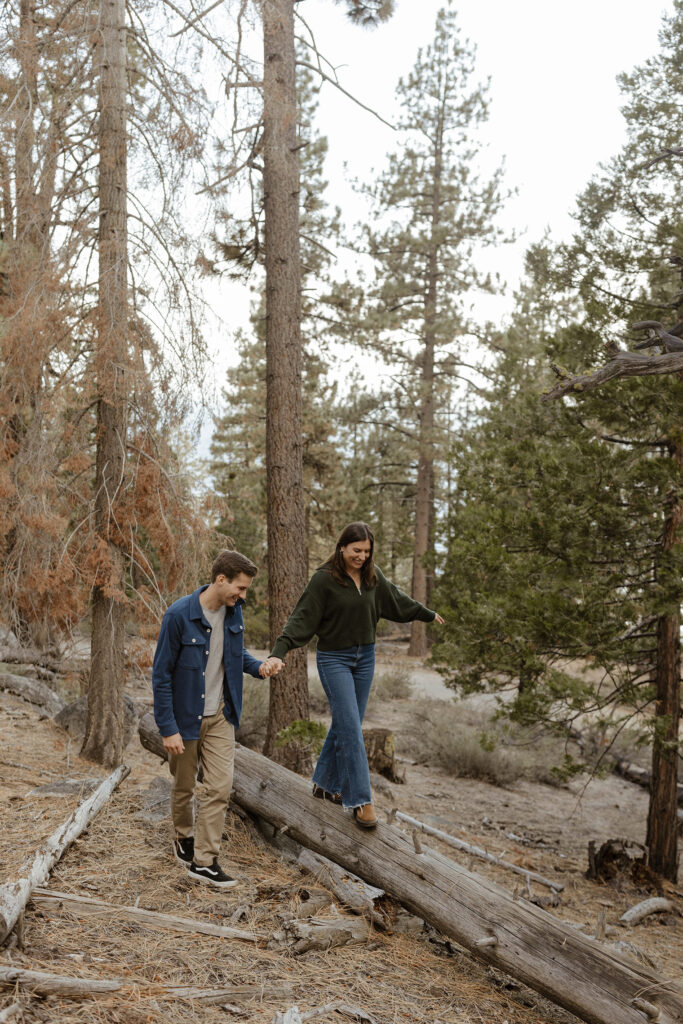 Woman walking across fallen tree while holding fiancé's hand and surrounded by pine trees and fallen pine needles in Lake Tahoe
