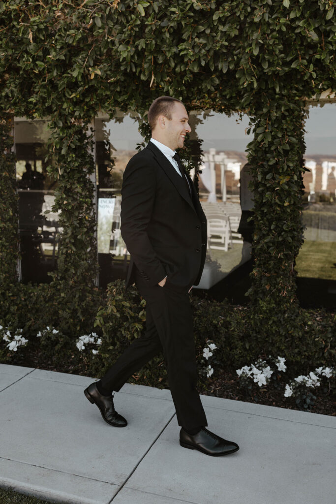 Wedding groom with hands in pockets walking along cement path in front of grass hedge with windows at Willow Heights mansion