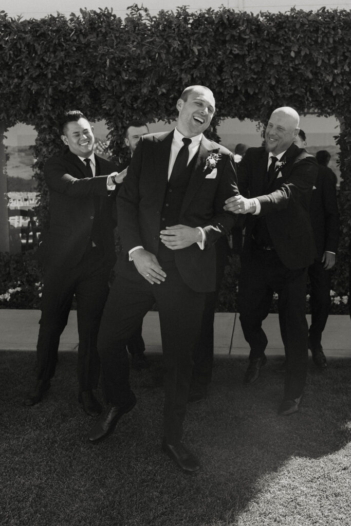 Wedding groom laughing while groomsmen grab and shove him playfully in front of grass hedge with windows at Willow Heights mansion