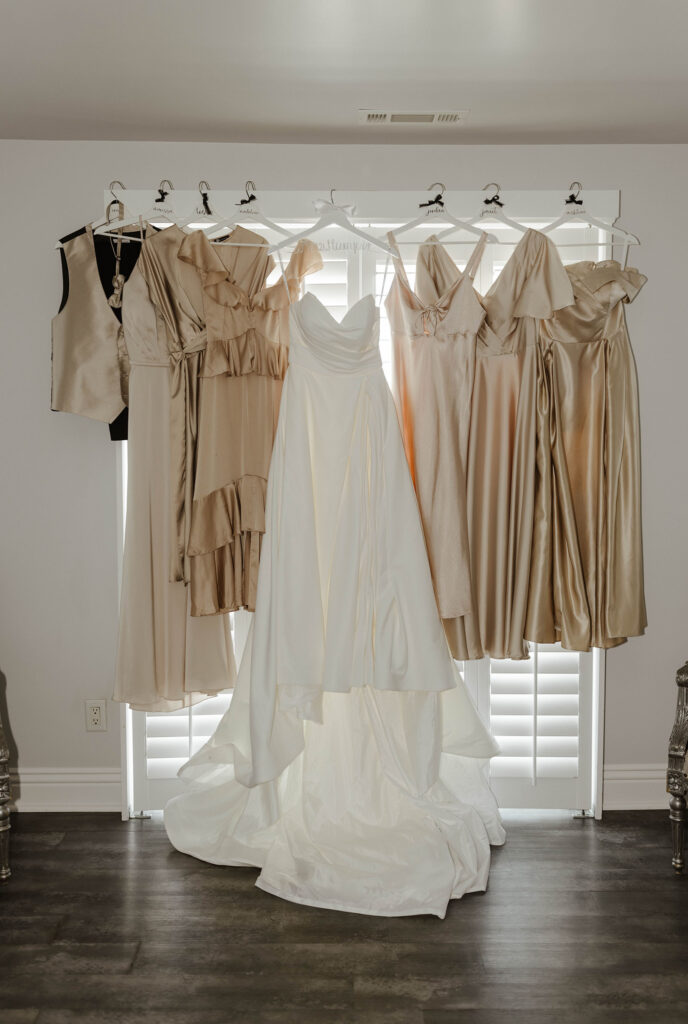 Bride's wedding dress and bridesmaids' dresses hanging in front of shuttered door with light coming through inside at Willow Heights mansion